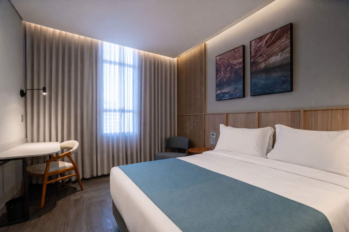 A hotel room with a bed, a desk, a chair, a window with curtains, and two wall-mounted paintings.