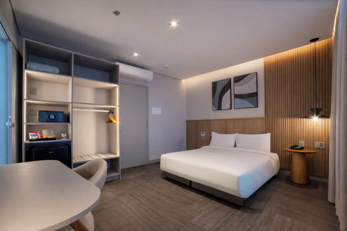 A modern hotel room with a bed, a desk, a chair, a wardrobe, and a small table with a lamp.