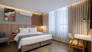 Une chambre d’hôtel moderne équipée d’un grand lit, d’un bureau, d’une chaise, d’un climatiseur fixé au mur et d’une fenêtre avec des rideaux.