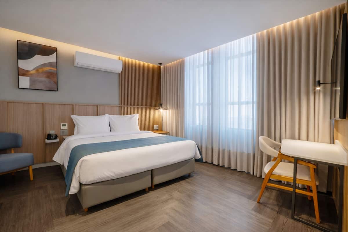 A modern hotel room with a large bed, a desk, a chair, a wall-mounted air conditioner, and a window with curtains.