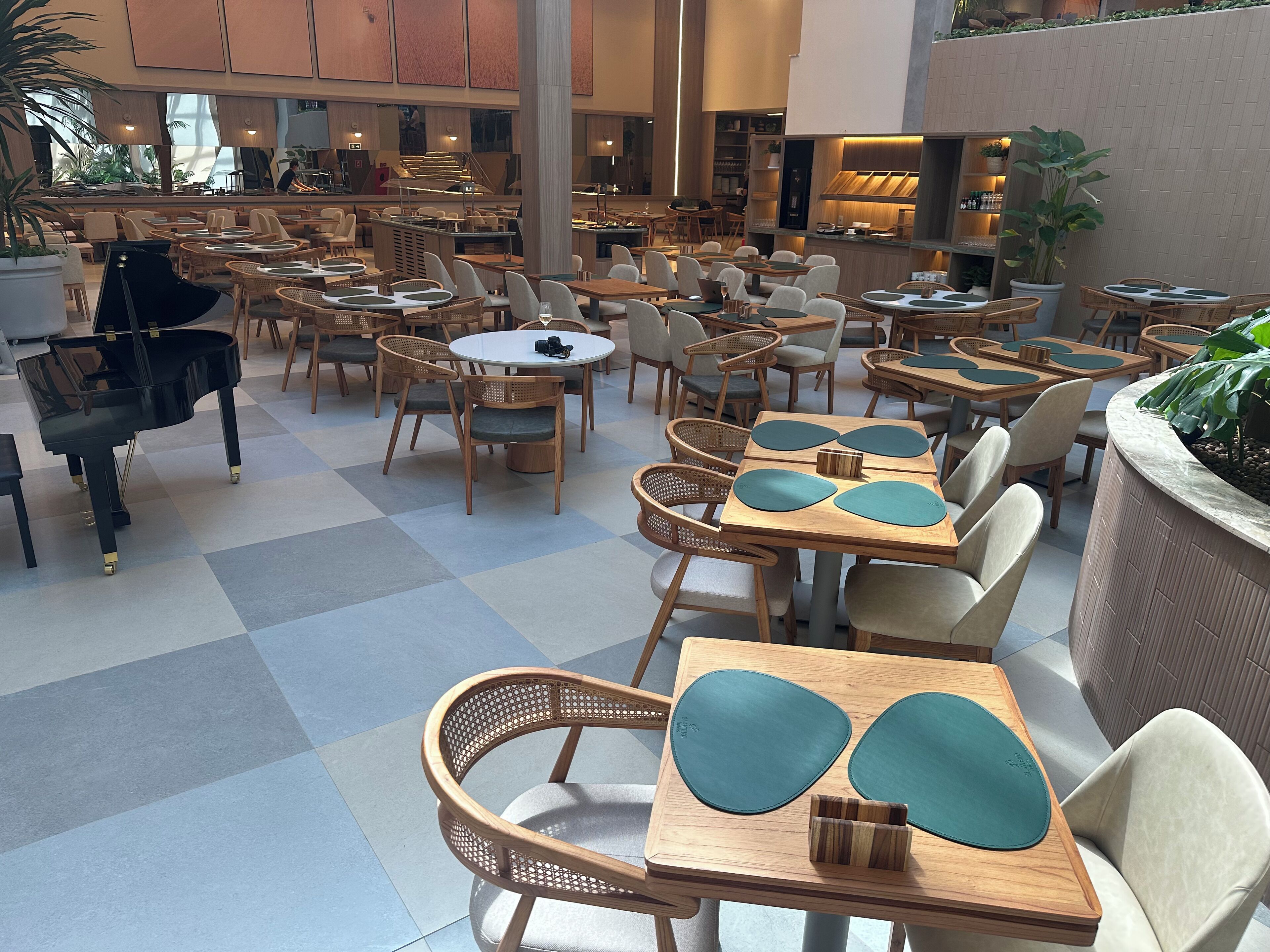 A spacious indoor dining area with multiple tables and chairs, a grand piano, and a variety of plants and shelves with items.