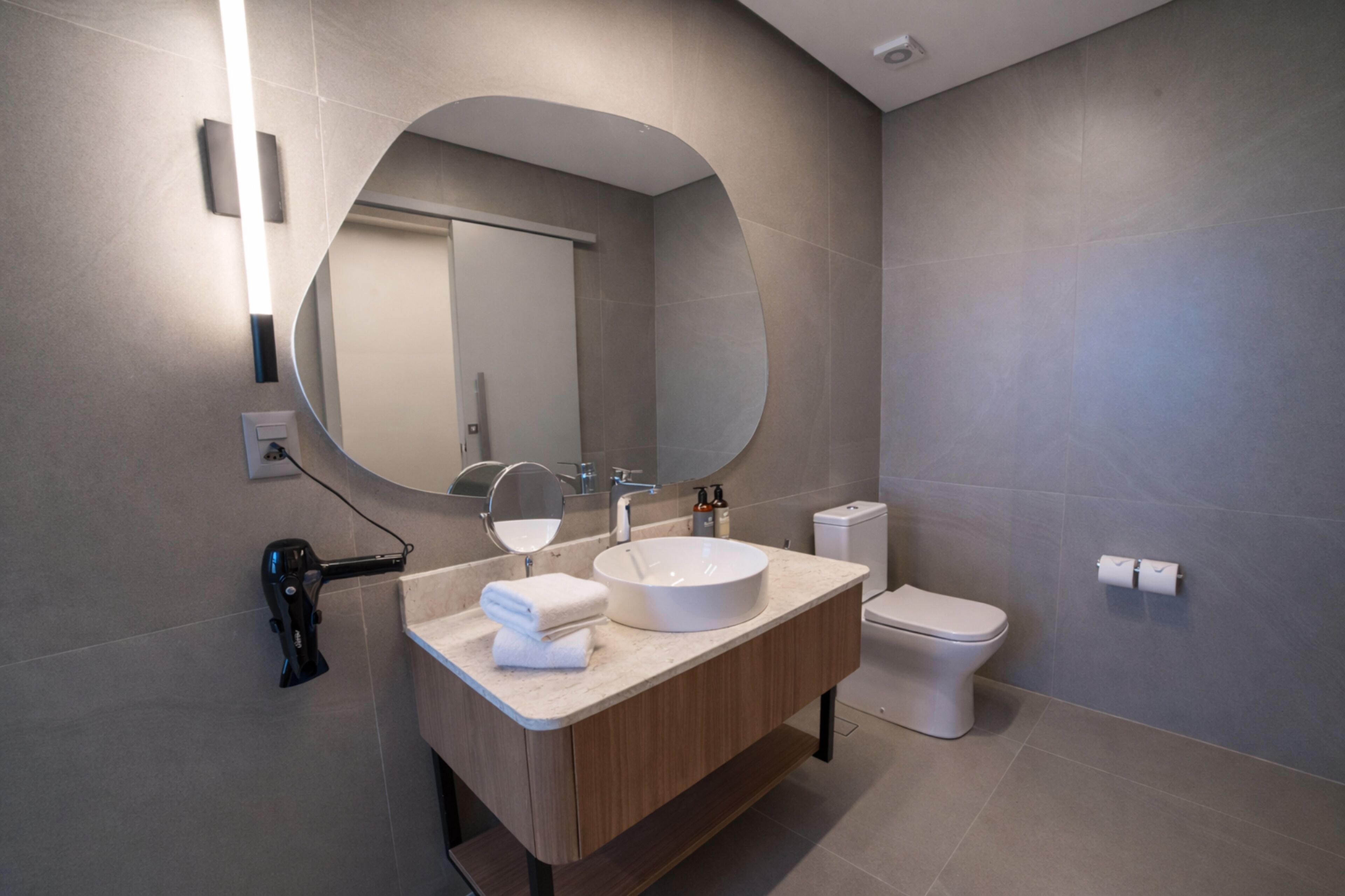 Un baño moderno con un espejo redondo, un lavamanos, un inodoro y un secador de pelo sobre el mostrador.