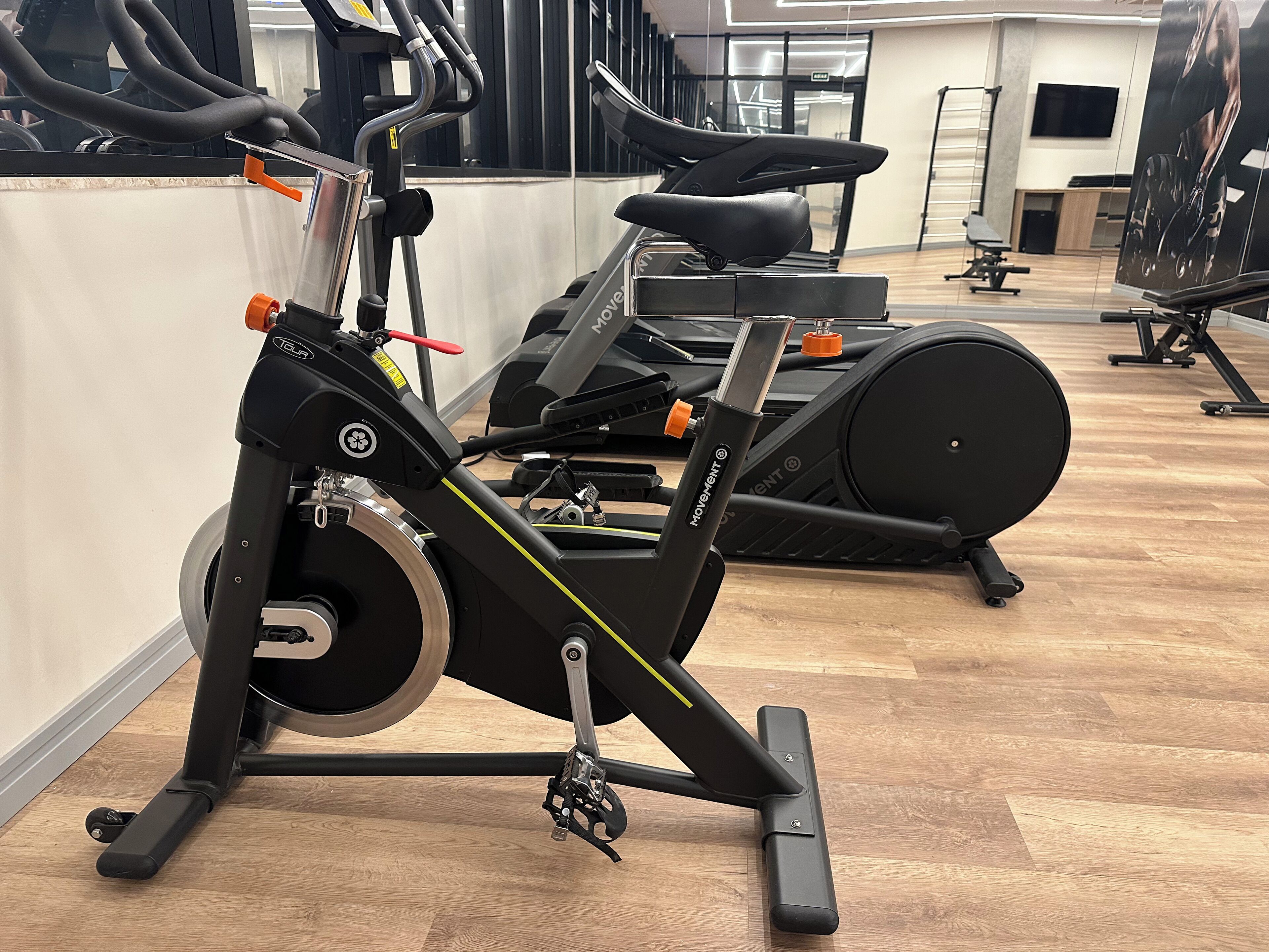 A stationary bike in a gym with a wooden floor, a wall-mounted TV, and other exercise equipment in the background.