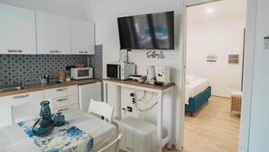 A compact kitchen with white cabinets, a microwave, and a coffee machine. A TV is mounted above the counter, and a bed is visible in the adjacent room. - Euterpe Apartment - Harmony, Relaxation and Sea at the Abode of the Muses (Praia a Mare)