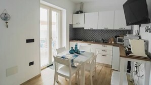 A compact kitchen with white cabinets, a dining table with chairs, a flat-screen TV mounted on the wall, and a window providing natural light. - Euterpe Apartment - Harmony, Relaxation and Sea at the Abode of the Muses (Praia a Mare)