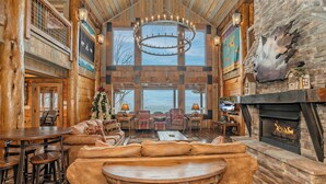 A spacious log cabin interior with a large fireplace, leather seating, and a high ceiling with exposed beams. - Mountain Cascades: Pool*Views*Theater*Games*Wifi*HotTub*Luxury (Sevierville)