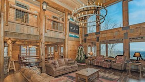 A spacious log cabin interior with a high ceiling, large windows, and a central fireplace. - Mountain Cascades: Pool*Views*Theater*Games*Wifi*HotTub*Luxury (Sevierville)