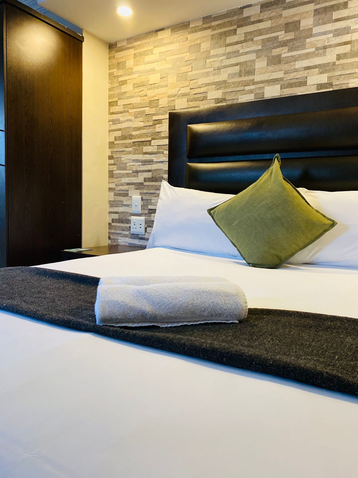 A hotel room with a bed, a headboard, a wall with a stone pattern, and a wooden wardrobe.