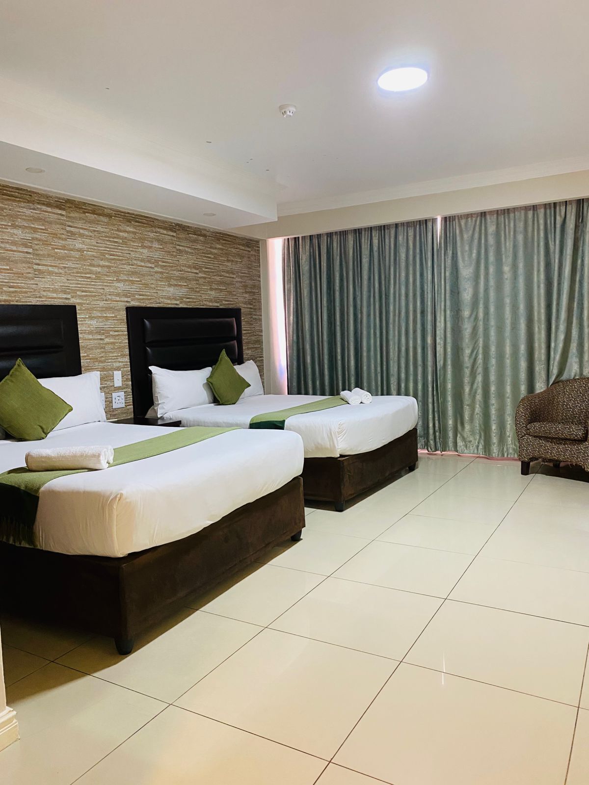 A hotel room with two beds, a large window with curtains, a patterned armchair, and a tiled floor.