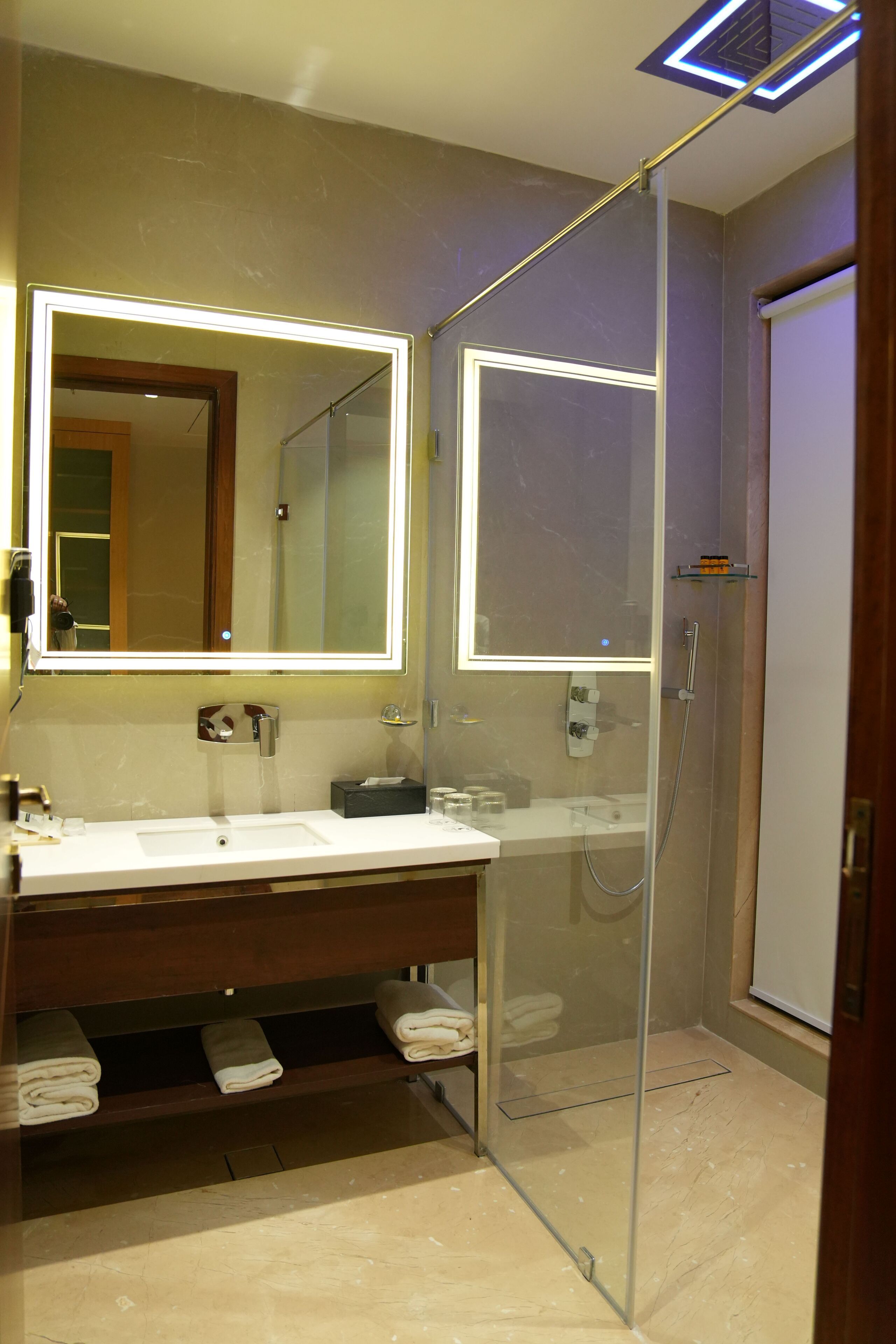 Ein modernes Badezimmer mit einer Duschkabine aus Glas, einem großen Spiegel mit LED-Beleuchtung und einem Waschtisch mit Spüle und Stauraum für Handtücher.