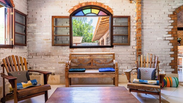 A rustic room with exposed brick walls, a wooden bench with cushions, two Adirondack chairs, and a large window with a view of a building and trees.