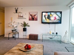 Un salón con televisor, una planta, un cuadro enmarco y una mesa con manzanas.