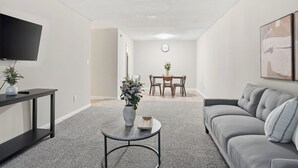 Un soggiorno moderno con un divano grigio, un tavolino da caffè, una TV a parete e, sullo sfondo, una zona pranzo.