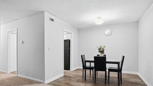 Een eetgedeelte met een tafel en stoelen, een klok aan de muur, en een deur die naar een andere kamer leidt.