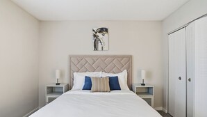 Een net opgemaakt bed met witte en blauwe kussens, een hoofdbord, nachtkastjes met lampen en een inloopkast met schuifdeuren.