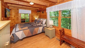 Quarto de cabana de madeira com cama, mesa de cabeceira, banco e televisão na parede.