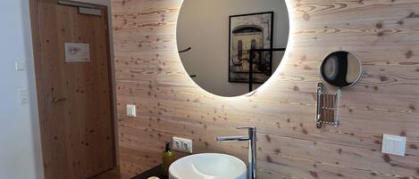 A modern bathroom with a wooden wall, a round mirror, a white sink, and a black countertop.