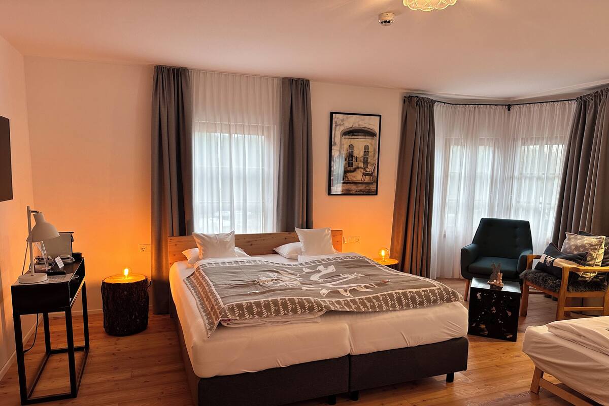 A well-lit bedroom with a large bed, a desk with a lamp, a chair, and a small table with a candle.