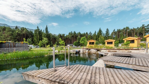 A wooden dock extending over a calm pond with a small waterfall, surrounded by lush greenery and forested hills. - Hotel Gerhardhof by ami (Wildermieming)