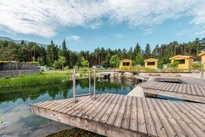 Eine Holzstege, der sich über einen ruhigen Teich mit einem kleinen Wasserfall erstreckt, umgeben von üppiger Vegetation und bewaldeten Hügeln.