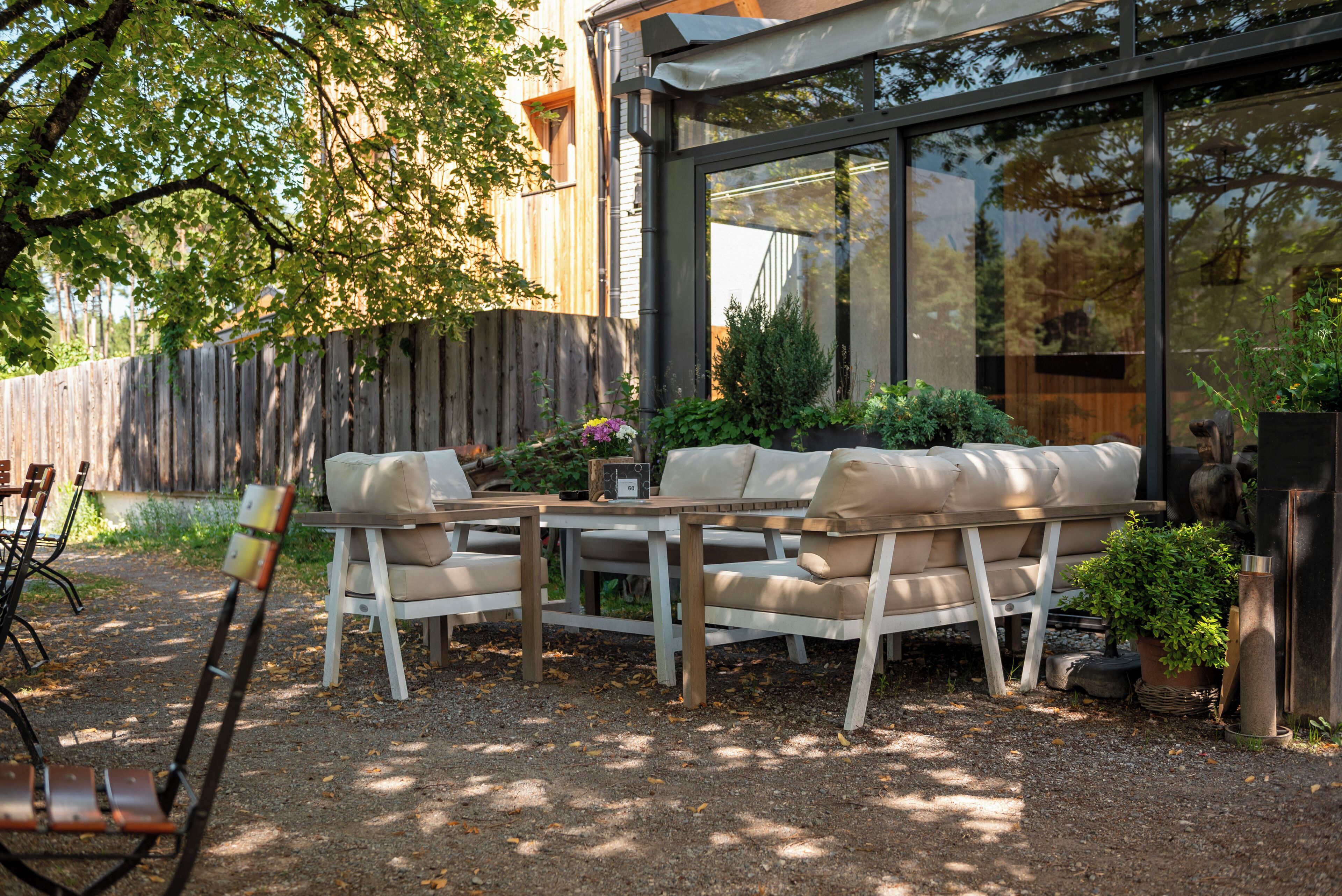 An outdoor seating area with a wooden table and beige cushioned chairs, surrounded by potted plants and a wooden fence.