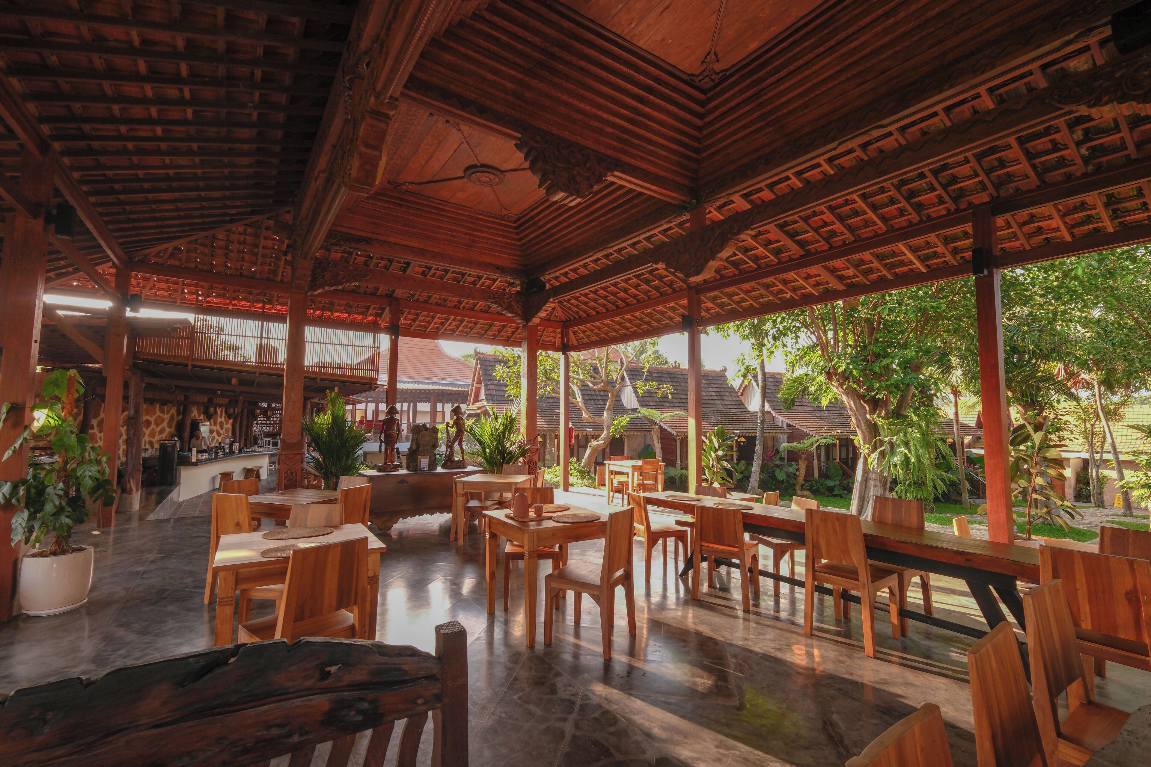 A covered outdoor dining area with wooden tables and chairs, potted plants, and a view of buildings in the background.