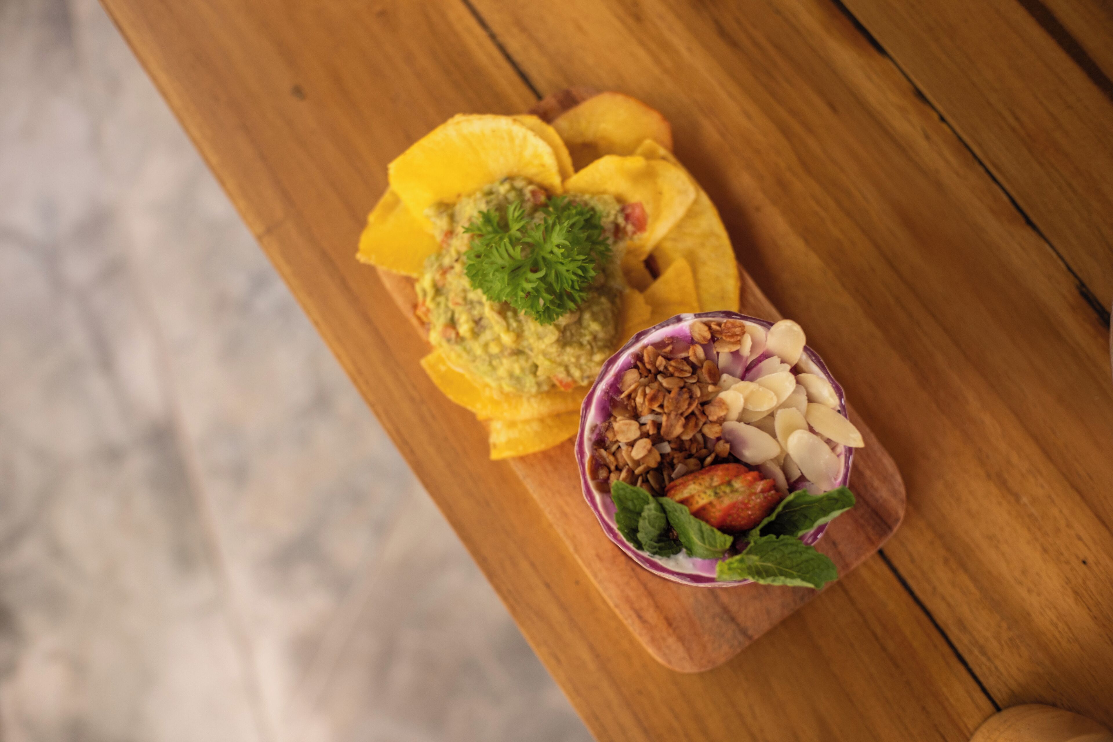 A wooden table with a bowl of guacamole and tortilla chips, and a bowl of fruit salad with almonds and mint.