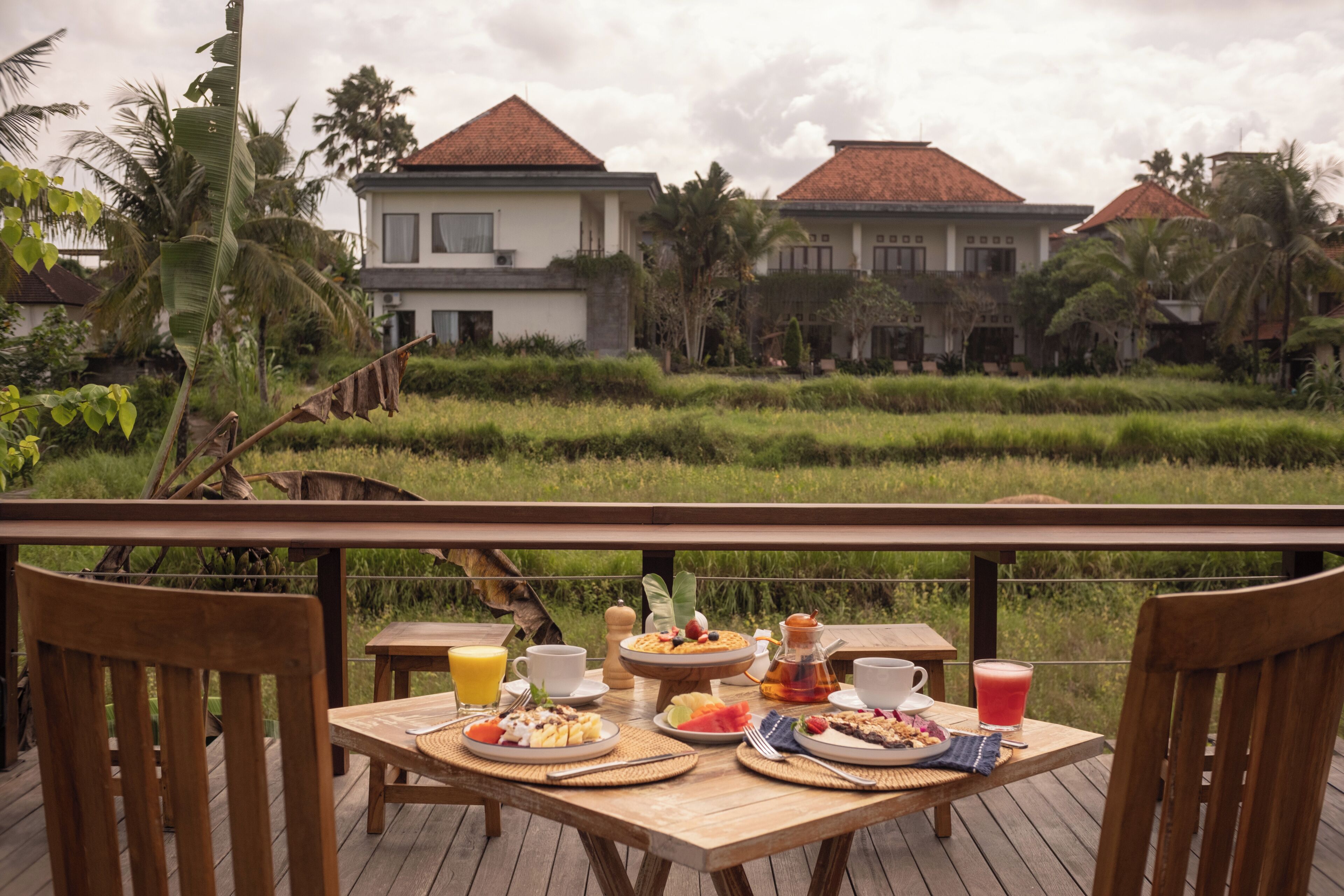 Een houten terras met een ontbijttafel, met uitzicht op een rijstveld en een huis met een rood dak.