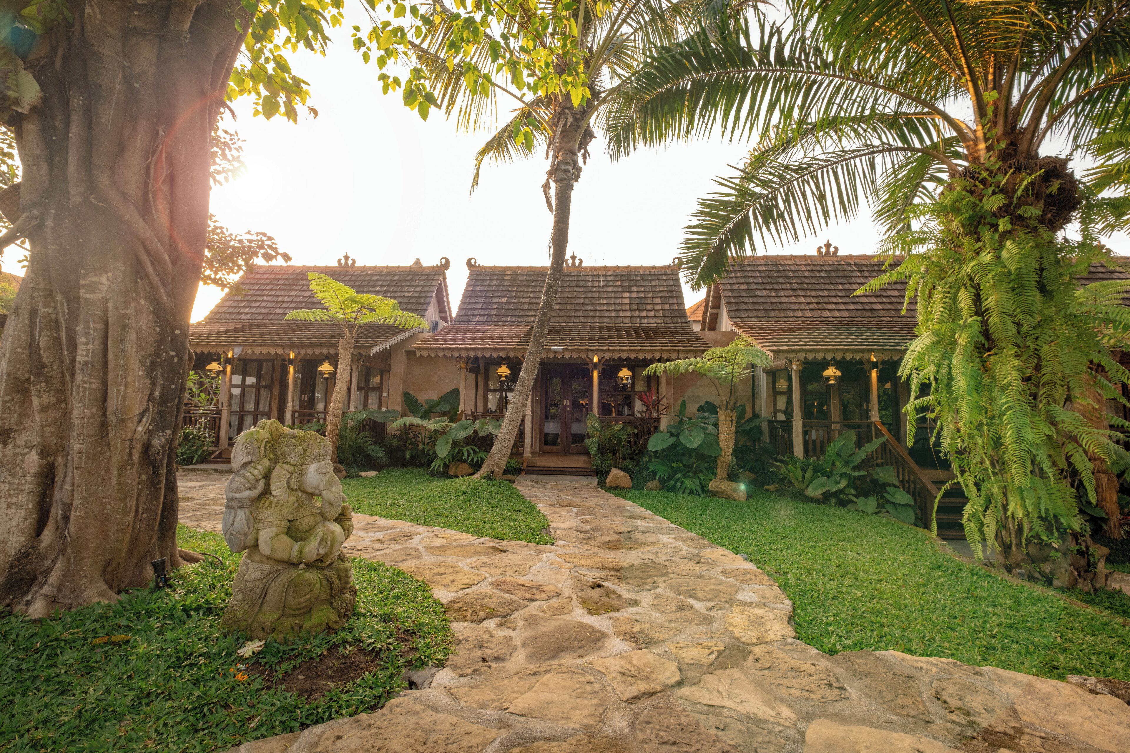 A traditional building with a stone pathway leading to the entrance, surrounded by lush greenery and palm trees.