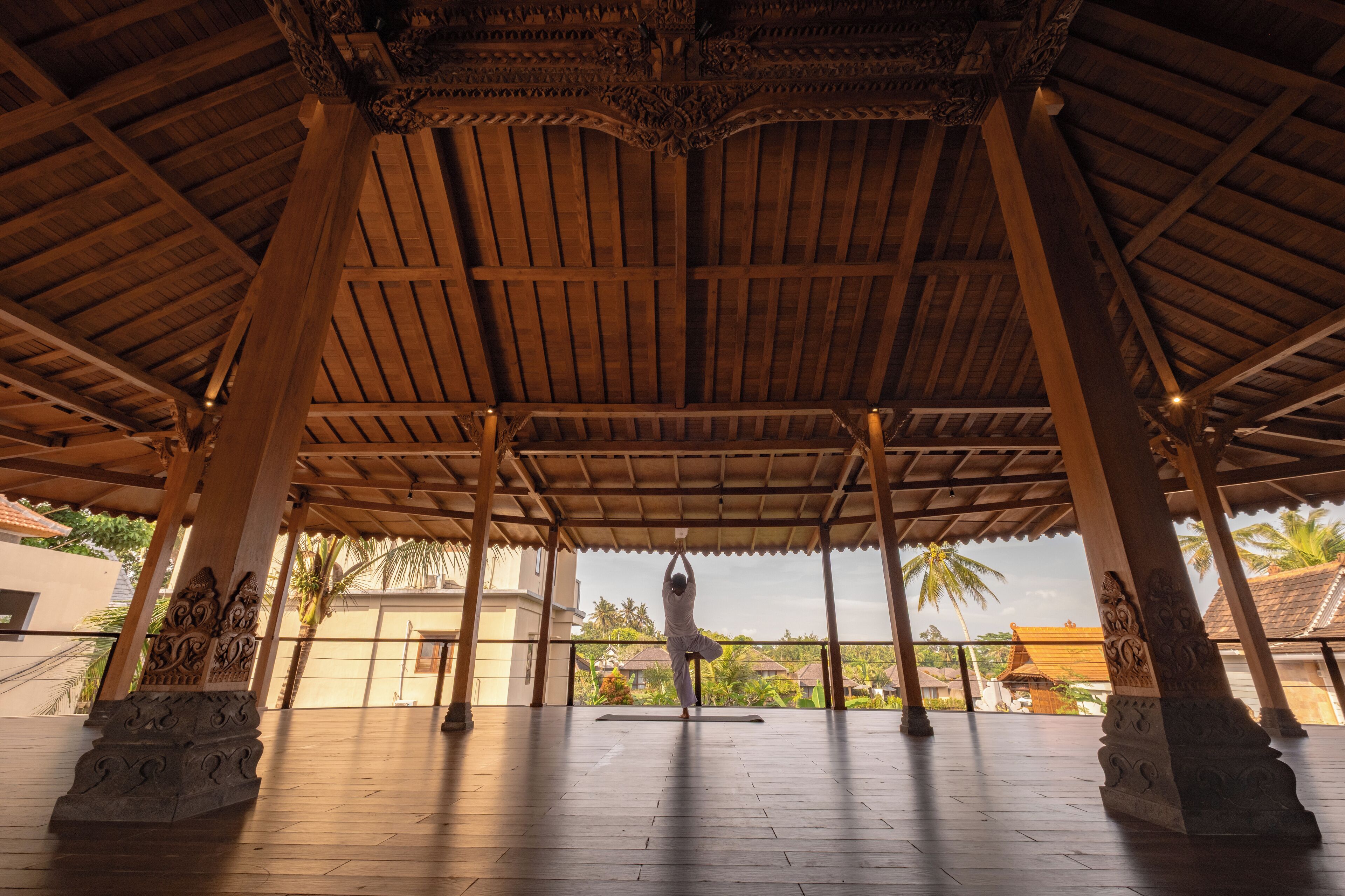 Een overdekte pergola met een houten plafond en zuilen, waar iemand een yoga-houding uitvoert.