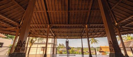 Een overdekte pergola met een houten plafond en zuilen, waar iemand een yoga-houding uitvoert.