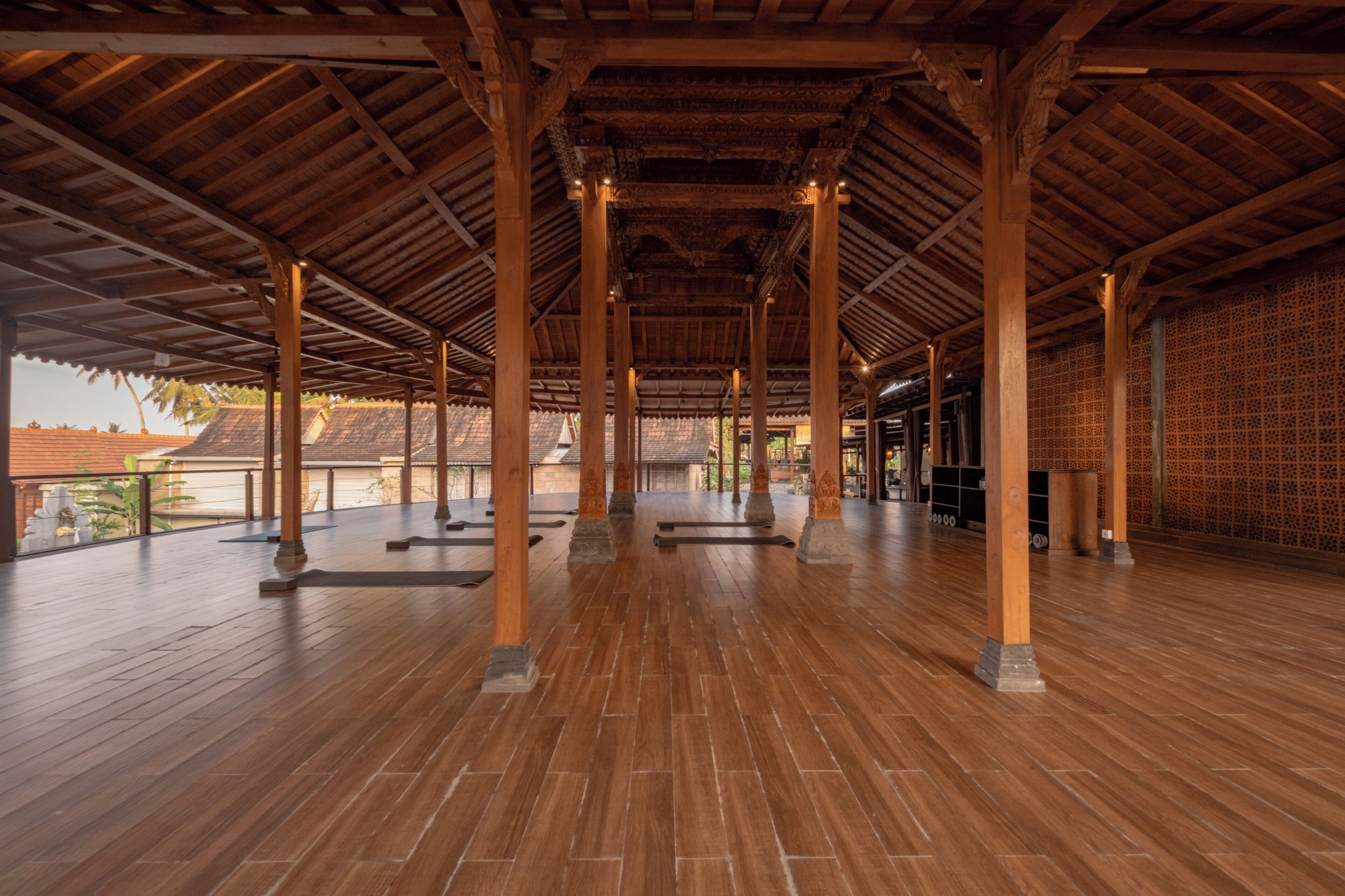 Un pavillon ouvert, doté d’un plancher en bois et de colonnes, offrant une vue sur des palmiers et un mur de briques en arrière-plan.