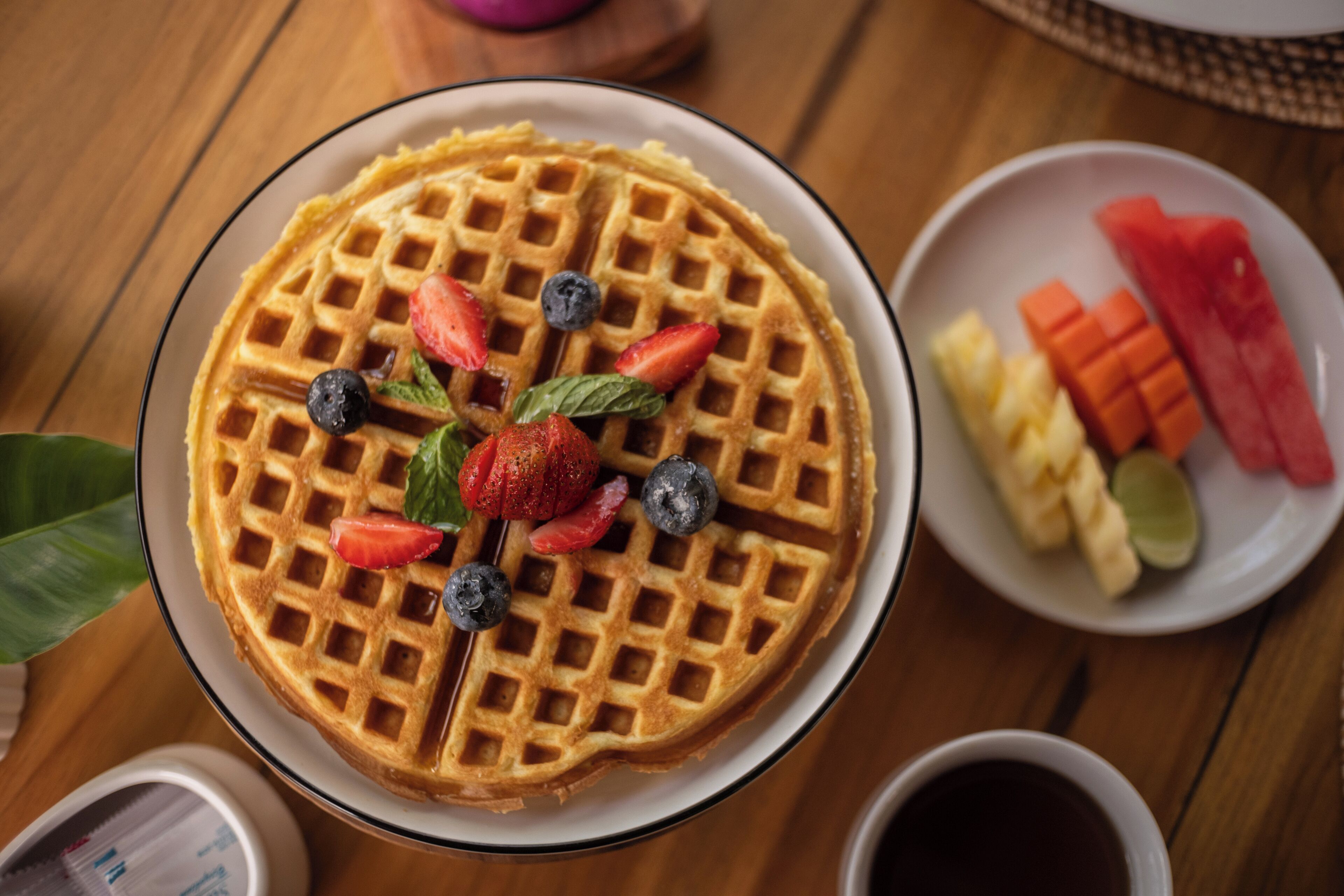 A waffle topped with strawberries and blueberries, accompanied by a plate of sliced fruits and a cup of coffee.
