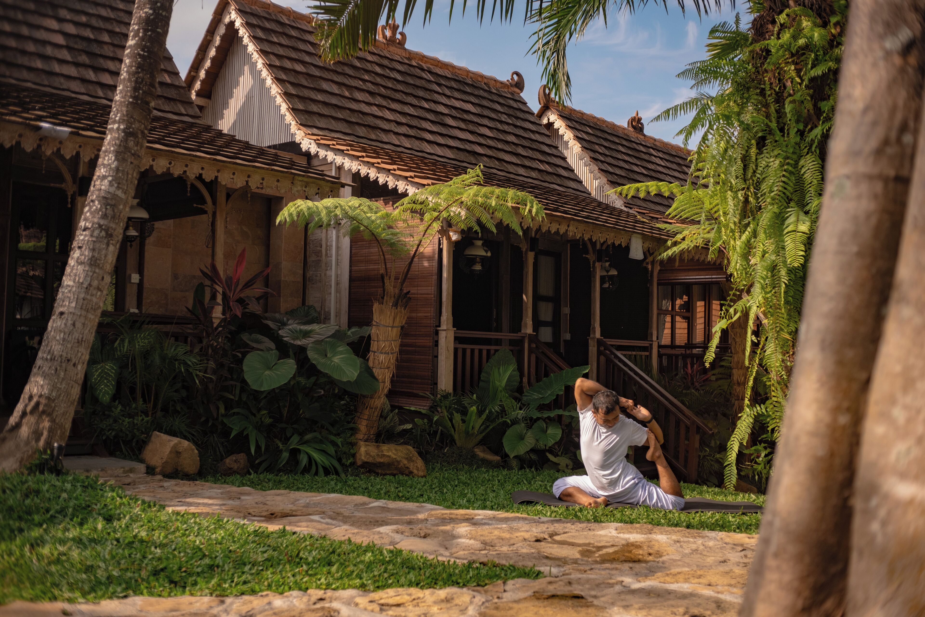 A traditional wooden house with a tiled roof, surrounded by lush greenery and palm trees.