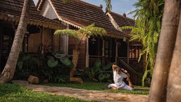 A traditional wooden house with a tiled roof, surrounded by lush greenery and palm trees.