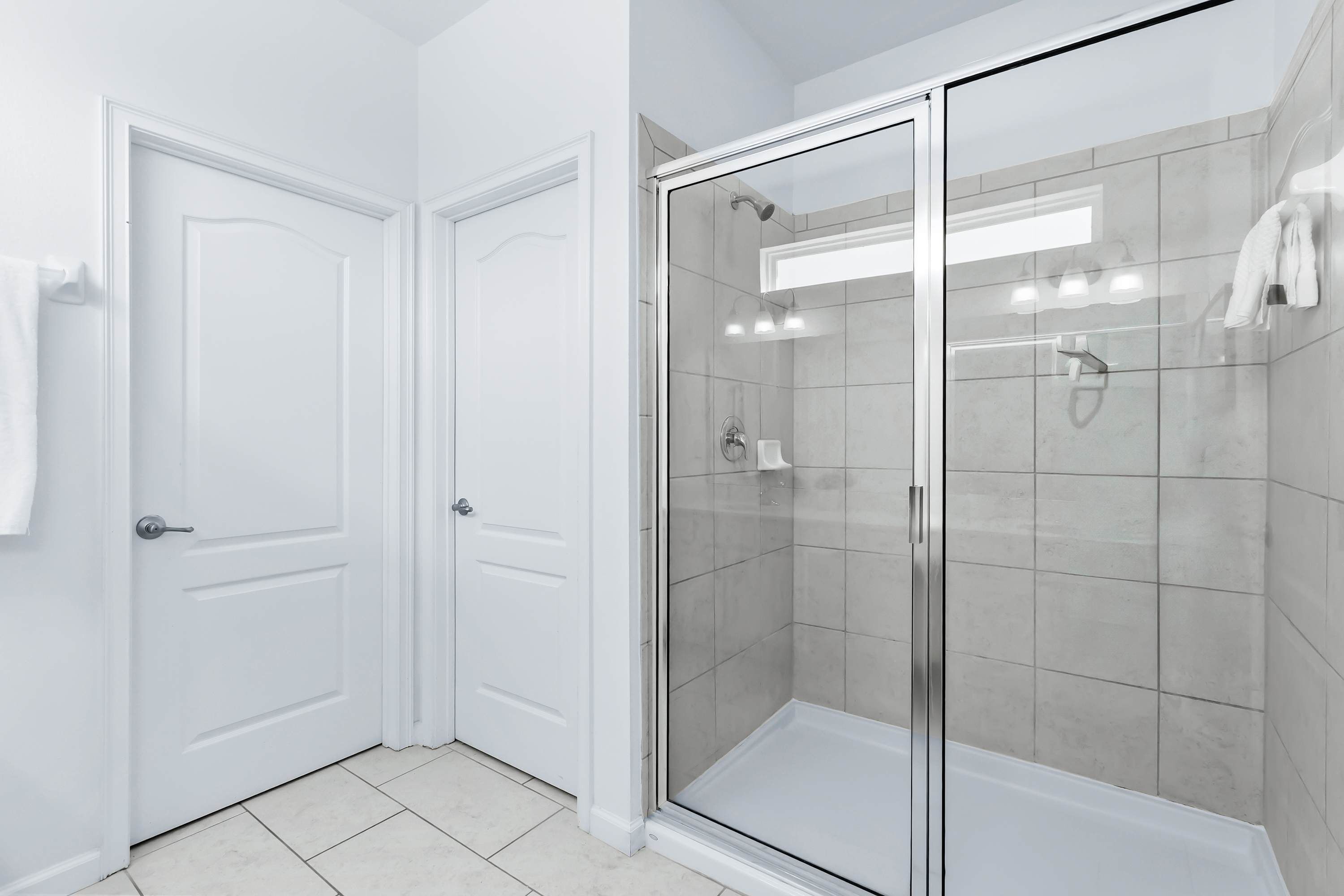 Une salle de bain moderne avec une douche à parois de verre, des murs recouverts de carreaux blancs et une porte sur laquelle est accrochée une serviette.