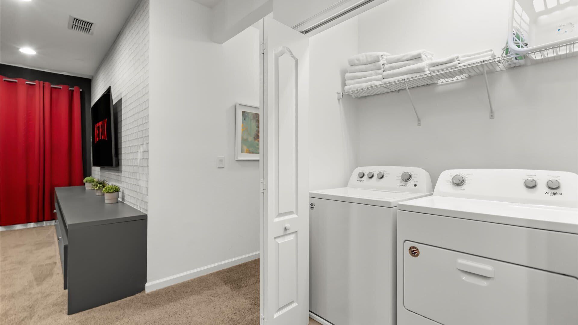 A laundry room with two Whirlpool washers and dryers, a white towel rack, and a red curtain.