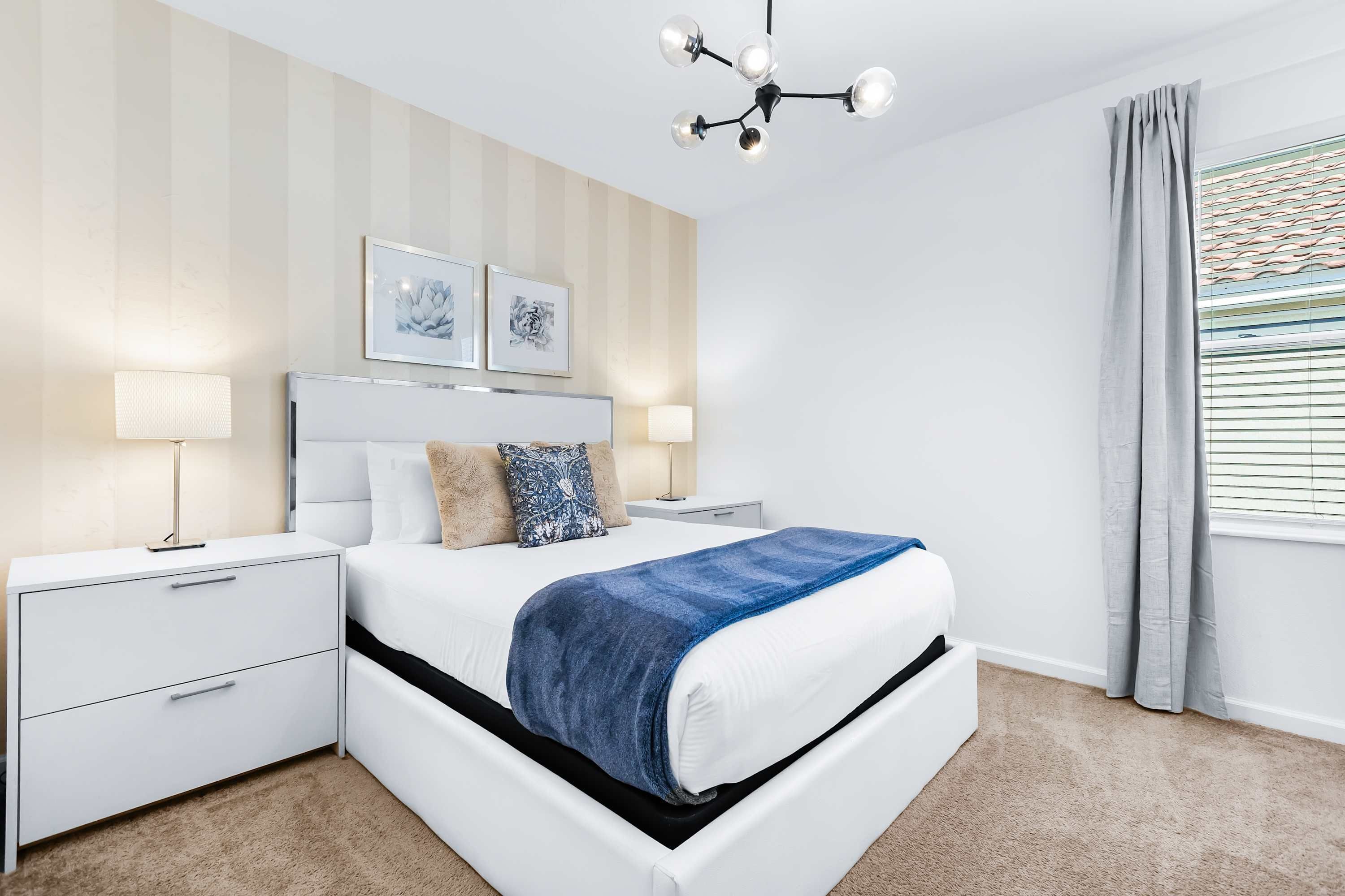 A modern bedroom with a white bed, two bedside tables, a chandelier, and a window with curtains.
