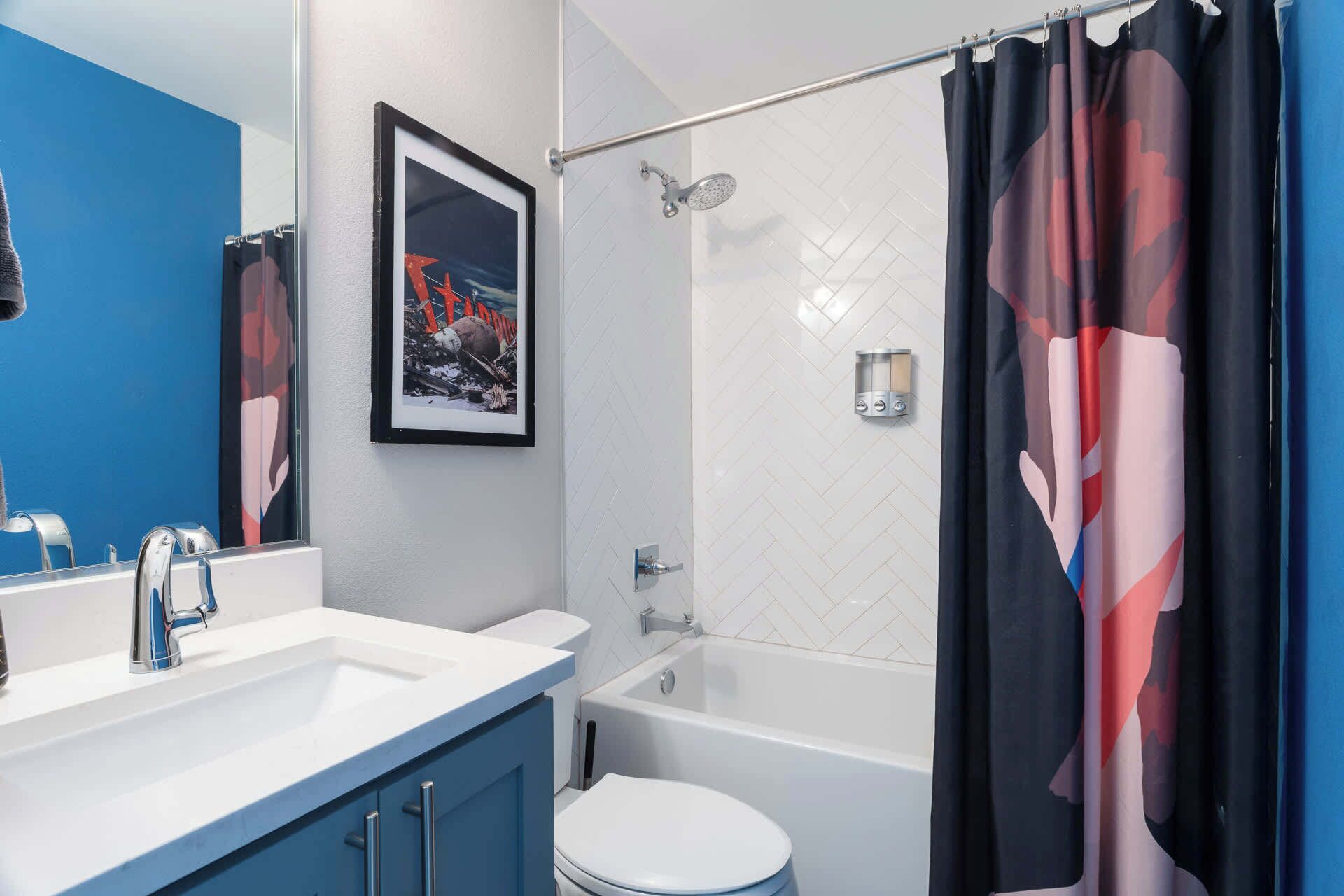 A bathroom with a white sink, blue cabinet, framed artwork, a shower with a glass door, and a toilet.