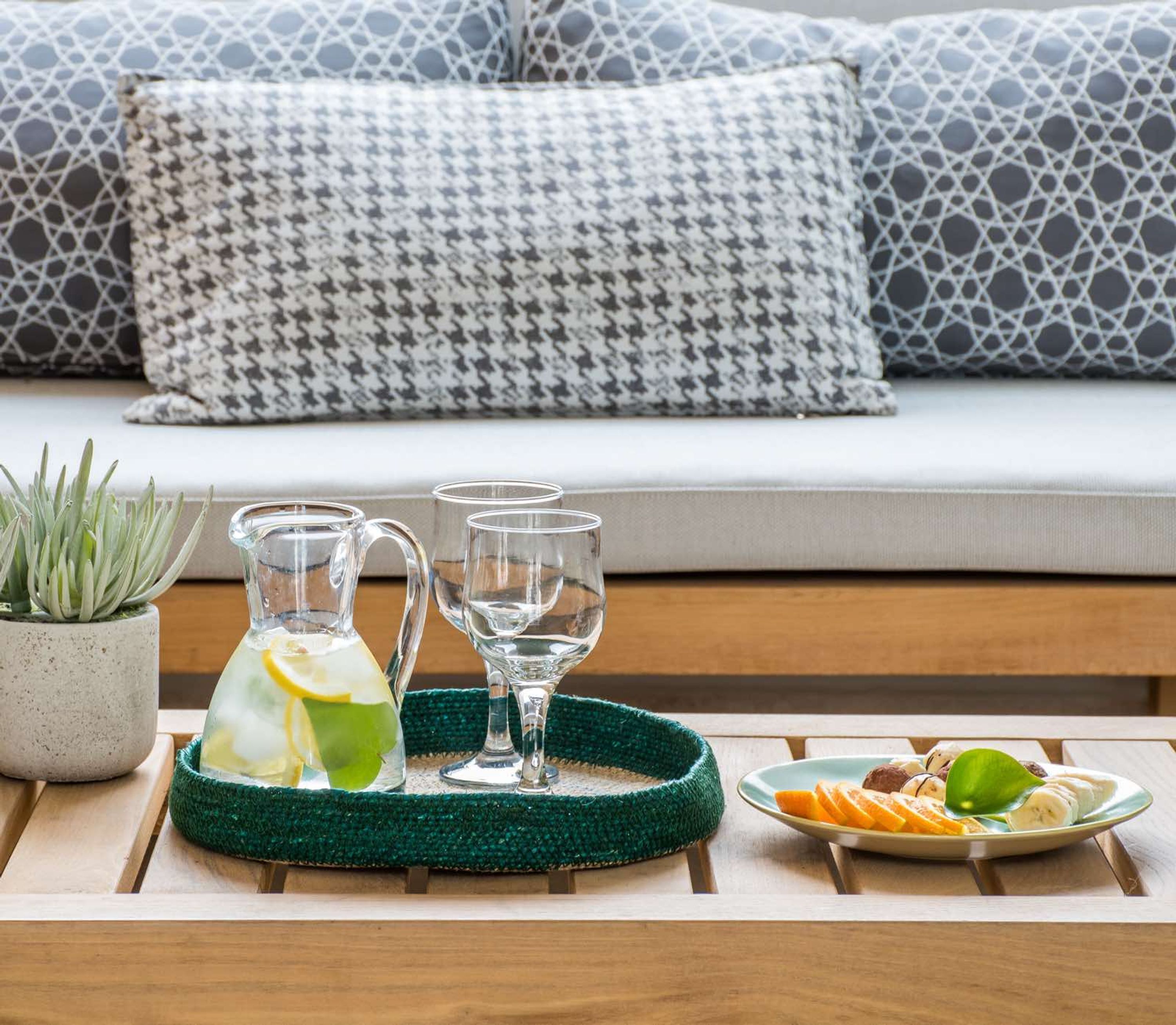 A tray with a pitcher and glasses on a wooden table, a plate with fruit, and a potted plant.