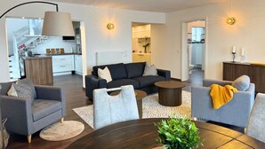 Un salon moderne avec un canapé gris foncé, deux fauteuils, une table basse et une table à manger. On aperçoit un escalier menant à l’étage supérieur, un coin cuisine et une salle de bain, cette dernière étant visible à travers une porte ouverte.