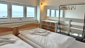 Une chambre d’hôtel avec deux lits, une tête de lit en bois, une commode blanche et une vue sur la mer grâce à de grandes fenêtres.