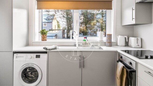 A modern kitchen with a built-in washing machine, a window with a view of the street, and a coffee maker. - The Den on Catto Drive (Peterhead)