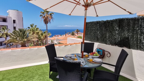 Ein Balkon mit Frühstückstisch, der Blick auf die Küste mit Palmen und einem Gebäude im Hintergrund bietet.