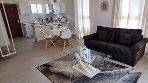 Ein modernes Wohnzimmer mit einem Glastisch, einem dunklen Sofa und einem Essbereich mit weißen Stühlen und einem Tisch.