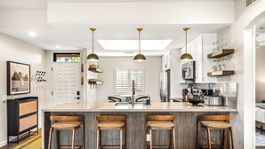 Une cuisine moderne dotée d’un îlot central, de tabourets de bar, de luminaires suspendus et offrant une vue sur l’extérieur grâce à une verrière.