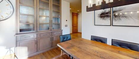 Un comedor con una mesa de madera, sillas, un armario con puertas de cristal y luces en la pared.