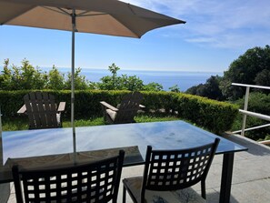 An outdoor patio with a glass table and chairs, a beige umbrella, and a view of the ocean. - VILLA AGA (Noli)