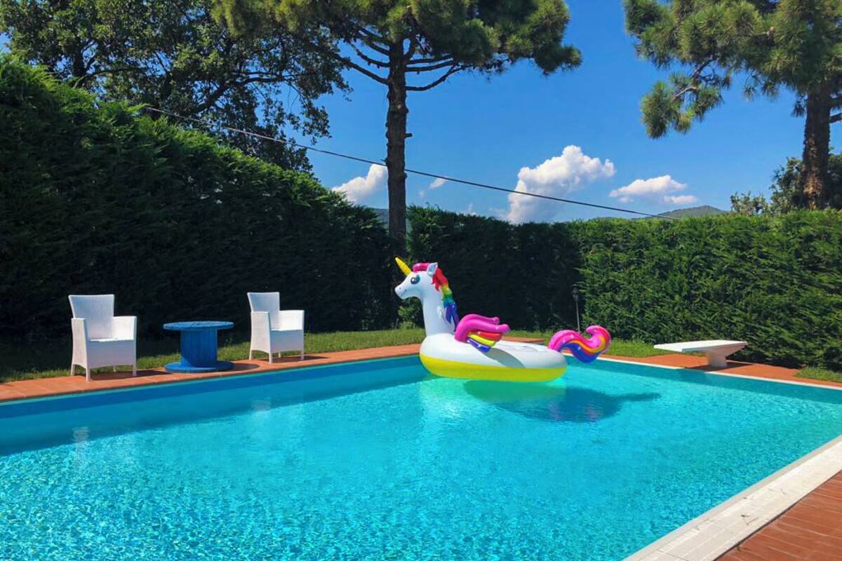 Una piscina con flotadores de unicornio inflables, sillas de descanso blancas y un suelo de ladrillo.