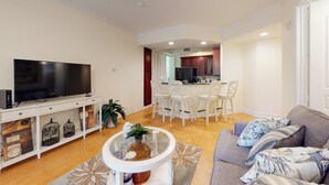 Una sala de estar moderna con un televisor de pantalla plana, un mueble blanco para televisión, una mesa de centro de vidrio y un sofá gris con cojines estampados.